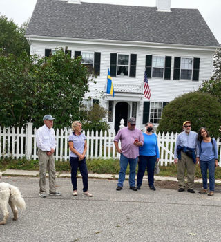 Group of people gathered in the street in front of an old, white house facing a tour guide who is talking