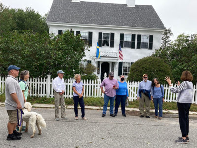 Group of people gathered in the street in front of an old, white house facing a tour guide who is talking