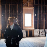 A woman, Pamela Cummings, standing in the foreground inside an old, uninsulated building with windows behind her. There are posters leaning up against the walls.