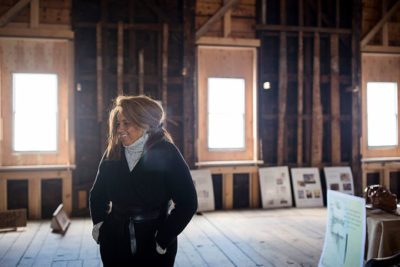 A woman, Pamela Cummings, standing in the foreground inside an old, uninsulated building with windows behind her. There are posters leaning up against the walls.